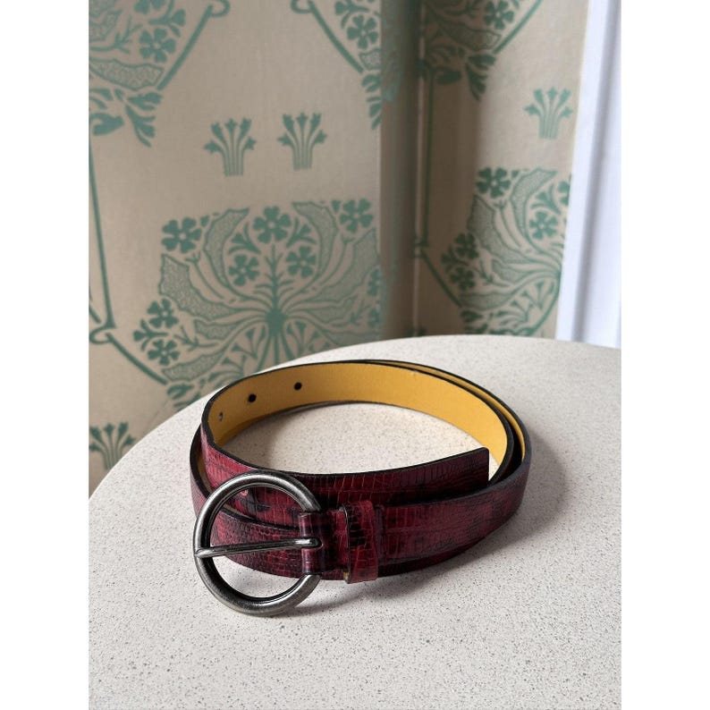 May include: A burgundy belt with a textured, reptile-like pattern. The belt has a large, round, silver-colored buckle and a yellow interior. The belt is displayed on a white surface.