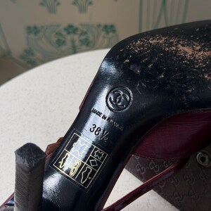 May include: A dark red high-heeled shoe with a black sole featuring the Chanel logo, the text Made in France, and the size 38.5. A square icon with symbols for leather and material composition is printed on the black heel area of the shoe.