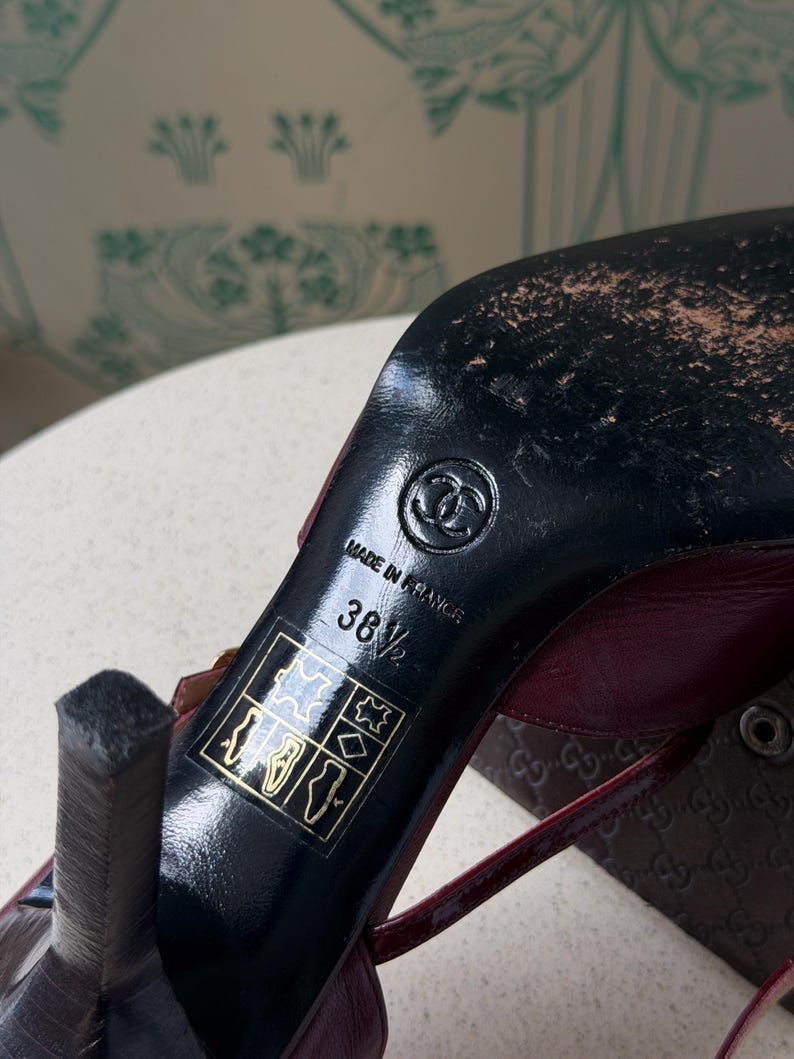 May include: A dark red high-heeled shoe with a black sole featuring the Chanel logo, the text Made in France, and the size 38.5. A square icon with symbols for leather and material composition is printed on the black heel area of the shoe.