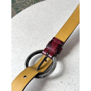 May include: A close-up of a yellow leather belt with a dark red, textured accent near the buckle. The belt features a silver-colored, circular buckle and a matching silver-colored belt loop. The belt is designed for waist sizes.