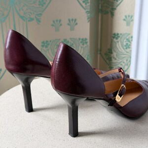 May include: A pair of dark burgundy leather Mary Jane pumps featuring a slim heel, a rounded toe, and a thin adjustable strap across the instep secured with a gold-toned metal buckle that displays a small logo emblem on the side.