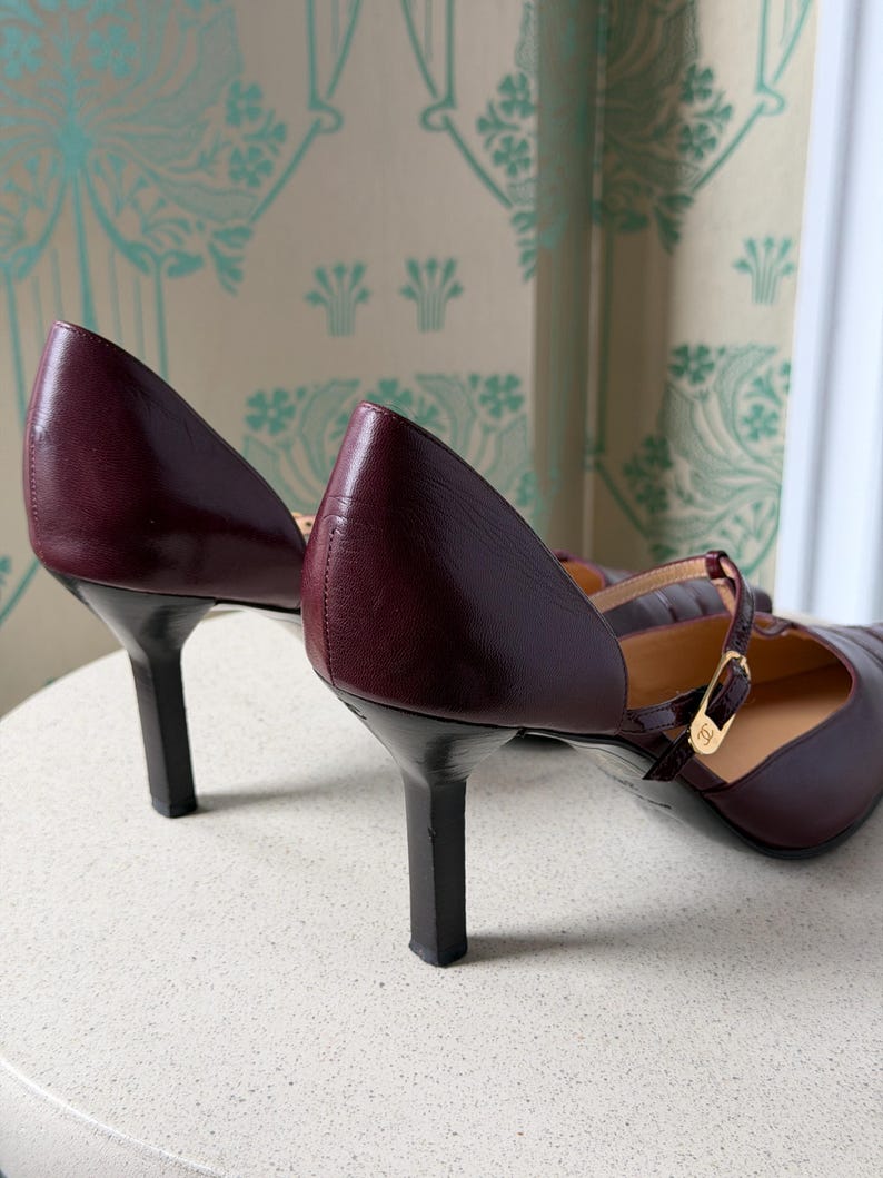 May include: A pair of dark burgundy leather Mary Jane pumps featuring a slim heel, a rounded toe, and a thin adjustable strap across the instep secured with a gold-toned metal buckle that displays a small logo emblem on the side.