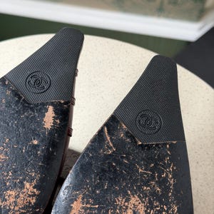 May include: The black soles of a pair of shoes featuring a pointed toe design. Each sole displays a textured black rubber insert at the tip, embossed with the interlocking CC Chanel logo. The remaining surface of the soles shows significant wear.