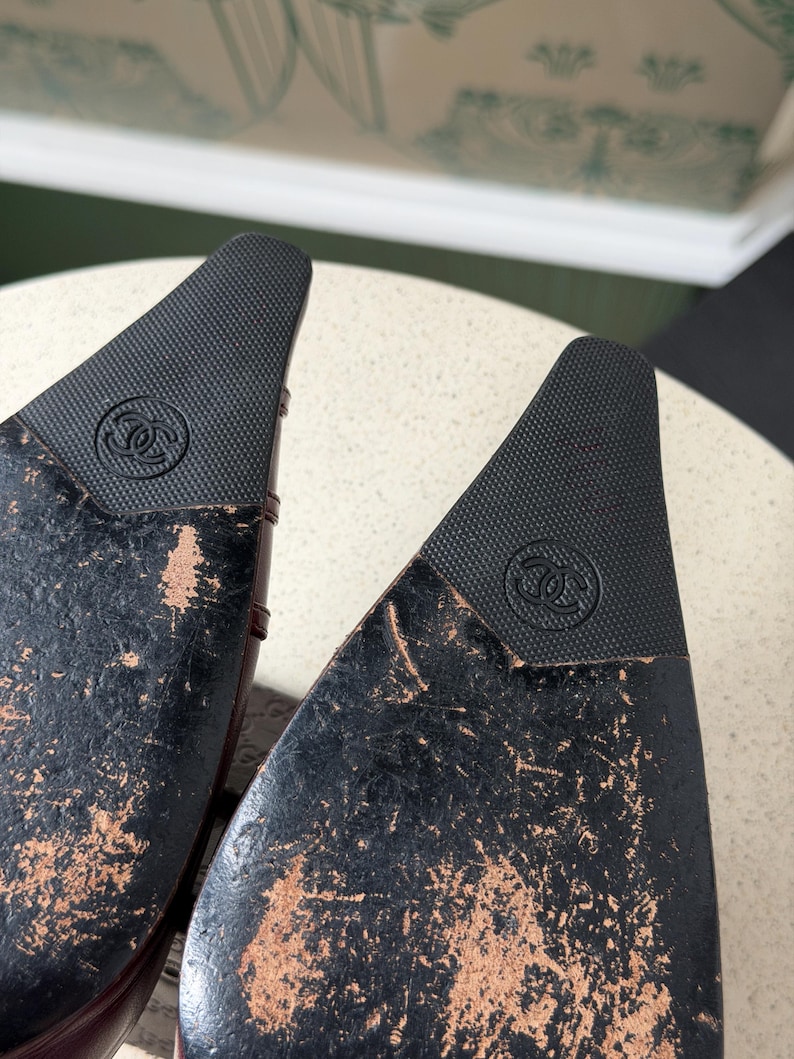 May include: The black soles of a pair of shoes featuring a pointed toe design. Each sole displays a textured black rubber insert at the tip, embossed with the interlocking CC Chanel logo. The remaining surface of the soles shows significant wear.