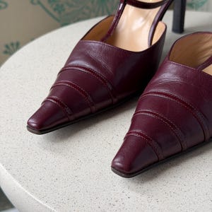 May include: A pair of burgundy leather pointed-toe pumps featuring a T-strap design with a buckle closure and a high heel. The shoes have a layered, ribbed texture on the toe box and the brand name CHANEL is visible on the inner sole.