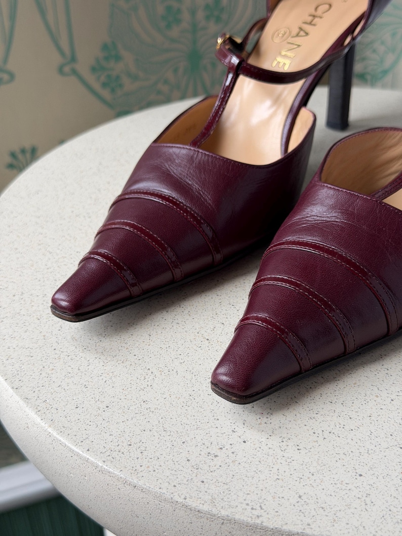 May include: A pair of burgundy leather pointed-toe pumps featuring a T-strap design with a buckle closure and a high heel. The shoes have a layered, ribbed texture on the toe box and the brand name CHANEL is visible on the inner sole.