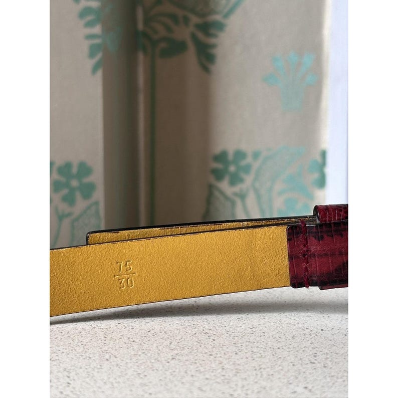 May include: Close-up of a leather belt with a burgundy textured finish and a gold-colored interior. The belt is marked with the size 75/30, indicating a 30-inch waist size. The background is a light-colored surface.