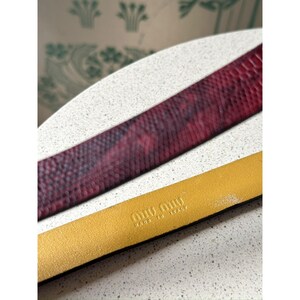 May include: A close-up of two leather belts. The top belt is a deep red with a snakeskin pattern. The bottom belt is a mustard yellow color with the brand name "MIU MIU" and "MADE IN ITALY" embossed on it.