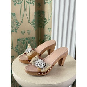May include: A pair of beige leather and cork platform sandals with a high heel. Each sandal features a large, sparkling crystal embellishment and a tan fabric accent. The heels are approximately 3 inches high.