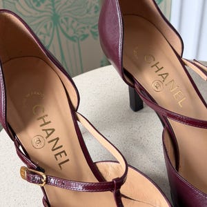May include: A pair of burgundy leather T-strap pumps with a pointed toe and a slim heel. The interior insole features the gold-stamped Chanel logo and the text Made in France. A thin adjustable ankle strap with a small gold-toned metal buckle.