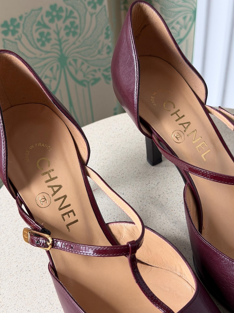 May include: A pair of burgundy leather T-strap pumps with a pointed toe and a slim heel. The interior insole features the gold-stamped Chanel logo and the text Made in France. A thin adjustable ankle strap with a small gold-toned metal buckle.