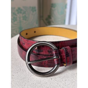 May include: A burgundy belt with a textured, reptile-like pattern. The belt features a large, circular, gunmetal-colored buckle. The interior of the belt is yellow. The belt is a fashion accessory.