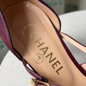 May include: A deep burgundy leather shoe featuring a tan leather insole with the gold-stamped text CHANEL and the CC logo. The shoe includes a thin burgundy strap with a gold-toned metal buckle that also displays the small CC logo.