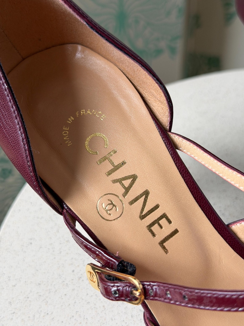 May include: A deep burgundy leather shoe featuring a tan leather insole with the gold-stamped text CHANEL and the CC logo. The shoe includes a thin burgundy strap with a gold-toned metal buckle that also displays the small CC logo.