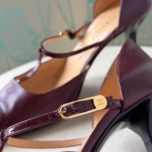 May include: A pair of deep burgundy leather T-strap heels featuring a gold-tone metal buckle with the Chanel logo. The shoes have a smooth leather finish, a pointed toe, and a slim heel. The interior lining is tan, and the brand name is visible.