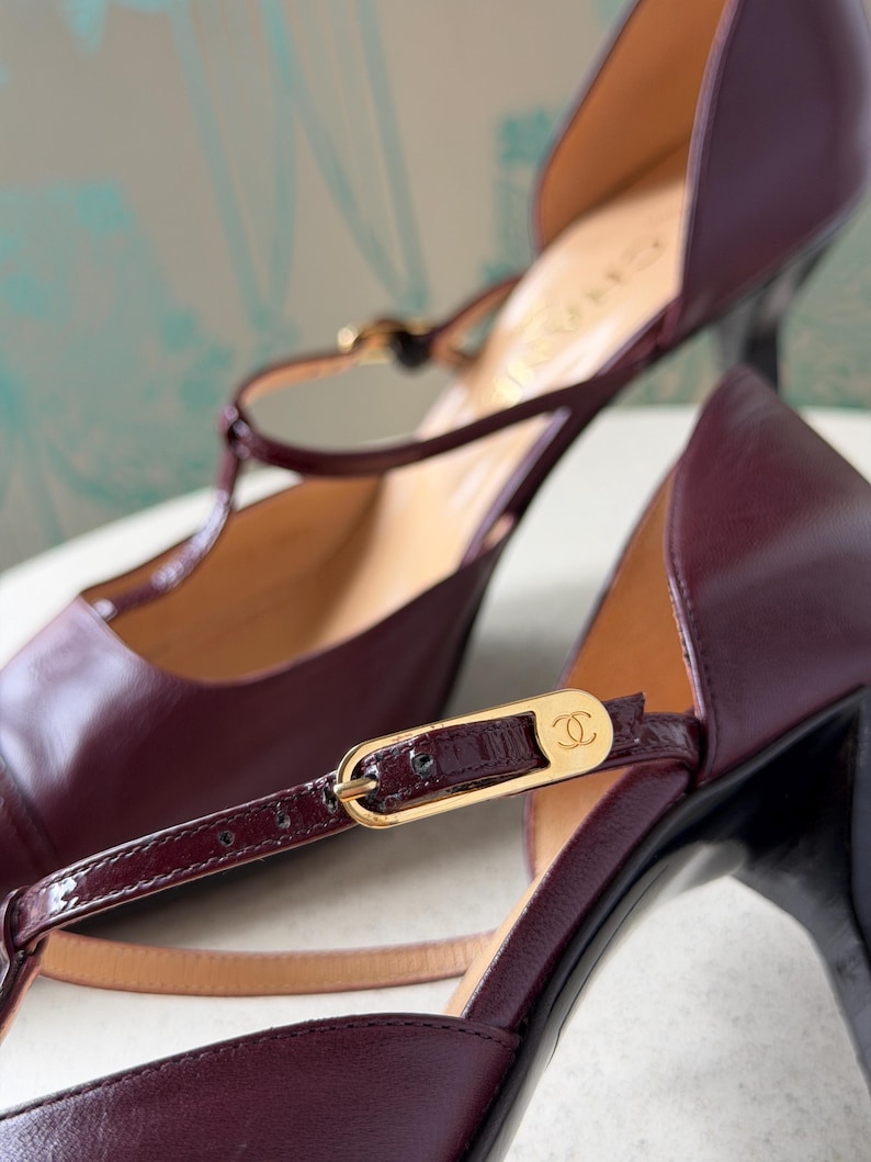 May include: A pair of deep burgundy leather T-strap heels featuring a gold-tone metal buckle with the Chanel logo. The shoes have a smooth leather finish, a pointed toe, and a slim heel. The interior lining is tan, and the brand name is visible.