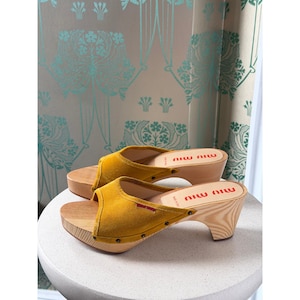 May include: A pair of mustard yellow suede mules with wooden soles and heels. The shoes feature a wide strap across the top, secured with small metal studs along the base. The brand name Miu Miu is printed in red on the light-colored wooden footbed.