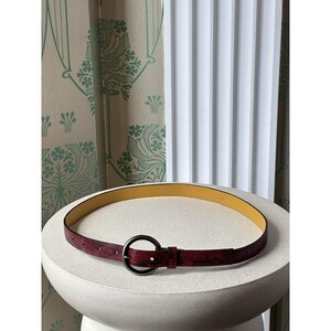 May include: A burgundy belt with a textured pattern and a black circular buckle. The interior of the belt is yellow. The belt is displayed on a white, rounded pedestal.