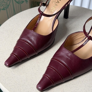 May include: A pair of burgundy leather T-strap pumps with a pointed toe, featuring horizontal stitched detailing across the vamp, a thin ankle strap with a buckle, and a slim heel. The interior insole is tan and displays the Chanel logo text.