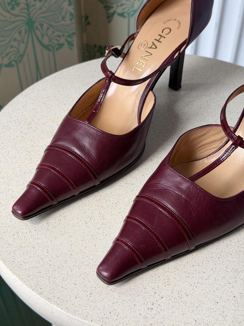 May include: A pair of burgundy leather T-strap pumps with a pointed toe, featuring horizontal stitched detailing across the vamp, a thin ankle strap with a buckle, and a slim heel. The interior insole is tan and displays the Chanel logo text.