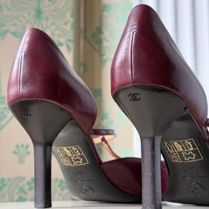 May include: A pair of burgundy leather pumps with thin heels, featuring the Chanel logo embossed on the heel and a gold-colored label on the sole. The shoes have a closed-toe design with a strap across the instep and a visible size marking of 38.5.