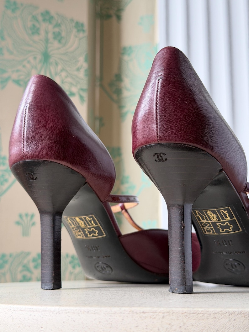 May include: A pair of burgundy leather pumps with thin heels, featuring the Chanel logo embossed on the heel and a gold-colored label on the sole. The shoes have a closed-toe design with a strap across the instep and a visible size marking of 38.5.