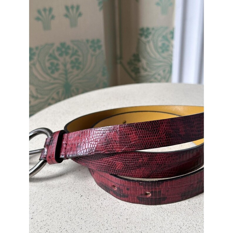 May include: A burgundy belt with a textured, reptile-like pattern. The belt has a silver-colored buckle and a yellow interior. The belt is coiled on a white surface, showcasing its design and details.