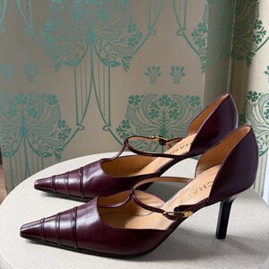 May include: A pair of dark burgundy leather T-strap pumps with a pointed toe, featuring horizontal stitched detailing on the toe box, a thin adjustable ankle strap with a gold-tone buckle, and a slim black heel. The interior sole is branded CHANEL.