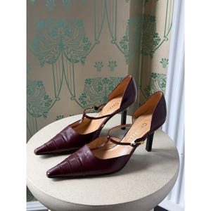 May include: A pair of dark burgundy leather pumps featuring a pointed toe, a T-strap design with a thin adjustable buckle, and a slim black heel. The interior sole is tan and clearly displays the brand name CHANEL printed in black lettering.