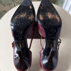 May include: A pair of burgundy leather slingback heels with pointed toes and black soles. The soles feature the Chanel logo, the text Made in France, size 38.5, and leather material icons. The heels are black with a small logo at the base.