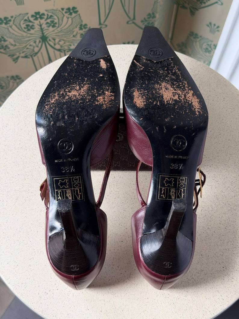 May include: A pair of burgundy leather slingback heels with pointed toes and black soles. The soles feature the Chanel logo, the text Made in France, size 38.5, and leather material icons. The heels are black with a small logo at the base.