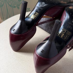 May include: A pair of burgundy leather high-heeled shoes with black heels. The soles are black and feature the Chanel logo, the text 'MADE IN ITALY', and the size 38.5. A small Chanel logo is embossed on the back of each black heel.