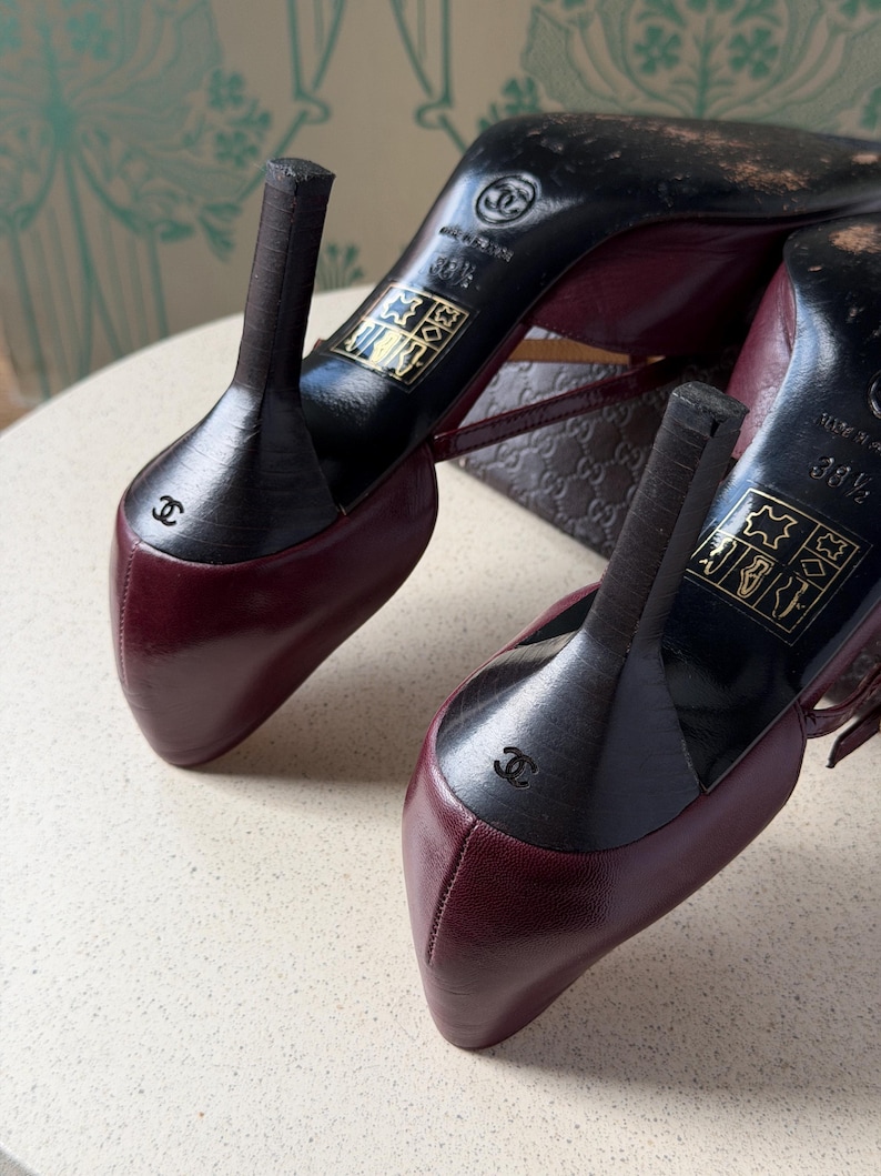 May include: A pair of burgundy leather high-heeled shoes with black heels. The soles are black and feature the Chanel logo, the text 'MADE IN ITALY', and the size 38.5. A small Chanel logo is embossed on the back of each black heel.