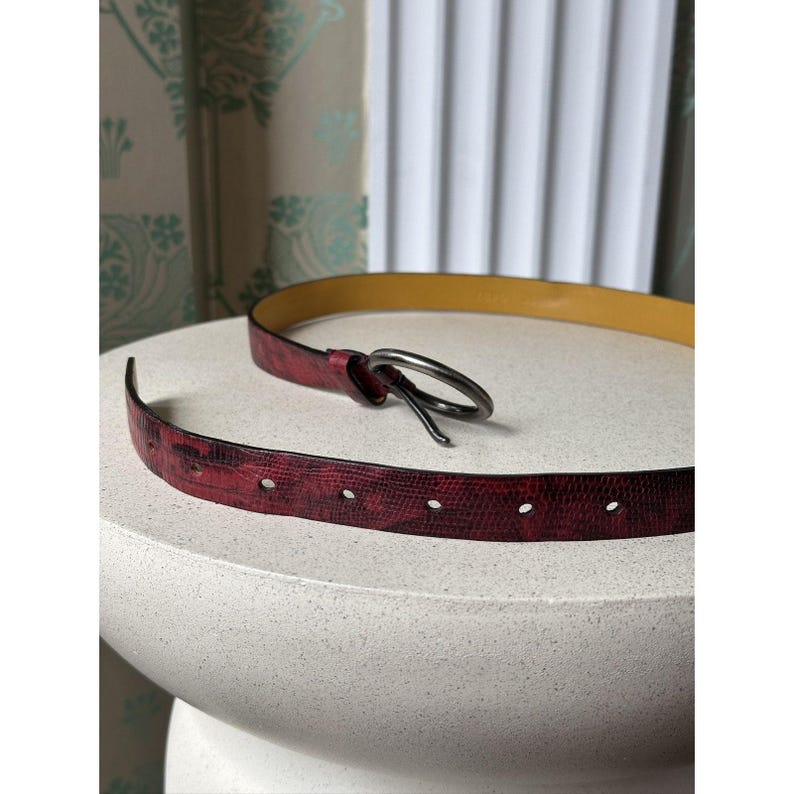May include: A red belt with a textured, snake-like pattern. The belt has a silver-colored, circular buckle and a yellow underside. The belt is lying on a white, rounded surface.