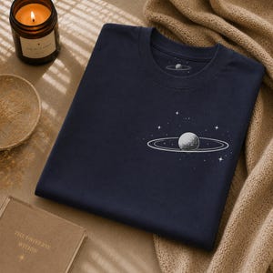 Pode incluir: Uma t-shirt azul-marinho com um gráfico branco de um planeta com anéis rodeado por pequenas estrelas no peito. Um gráfico de planeta mais pequeno a condizer está impresso na etiqueta interior do pescoço. T-shirt com gola redonda.