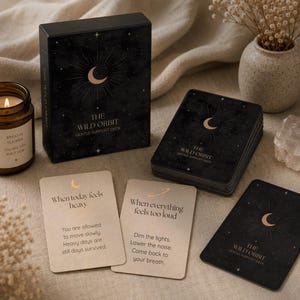 Pode incluir: Uma caixa preta e um baralho de cartas com a etiqueta "THE WILD ORBIT GENTLE SUPPORT DECK" com um design de meia-lua. Duas cartas estão viradas para cima, mostrando texto. Uma vela acesa e um pequeno vaso com flores secas também estão presentes.