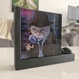 Op de afbeelding: Een ingelijst kunstwerk met een kat als detective, met een vergrootglas, een hoed en een trenchcoat. De achtergrond toont het opschrift "Milk Bar". Het zwarte frame heeft de initialen "AJ".