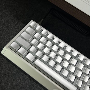 May include: A compact mechanical keyboard featuring a silver metallic frame and matching silver keycaps with black lettering. The keyboard includes a standard QWERTY layout with an ESC key, a long spacebar, and a black coiled cable connected at the top.