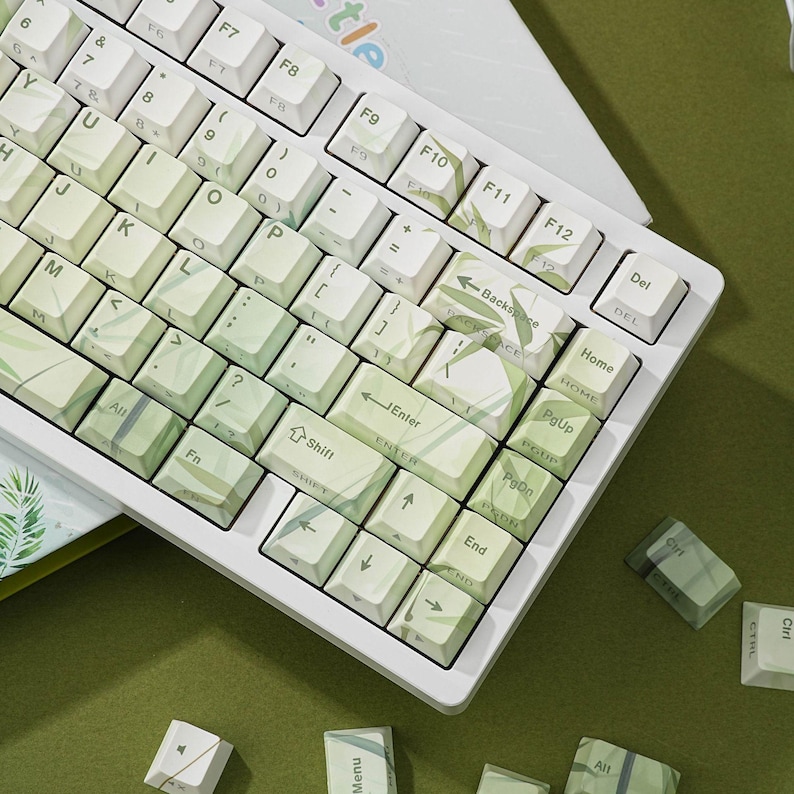 May include: White mechanical keyboard with green and white keycaps. The keycaps have a leaf pattern. The keyboard is on a green surface. Some extra keycaps are scattered around the keyboard.