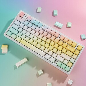May include: A white mechanical keyboard with pastel-colored keycaps arranged in a gradient from pink to yellow. Several loose keycaps are scattered around the keyboard on a colorful background.