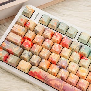May include: A white mechanical keyboard with custom keycaps. The keycaps feature a floral design in shades of orange, red, yellow, and green. The keyboard has standard keys, including function keys and a number pad.