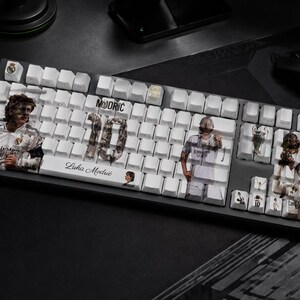 May include: A black mechanical keyboard with white keycaps featuring custom graphics of Luka Modrić, the number 10, and Real Madrid imagery. The spacebar displays the text Luka Modrić, while other keys show various player portraits and trophies.