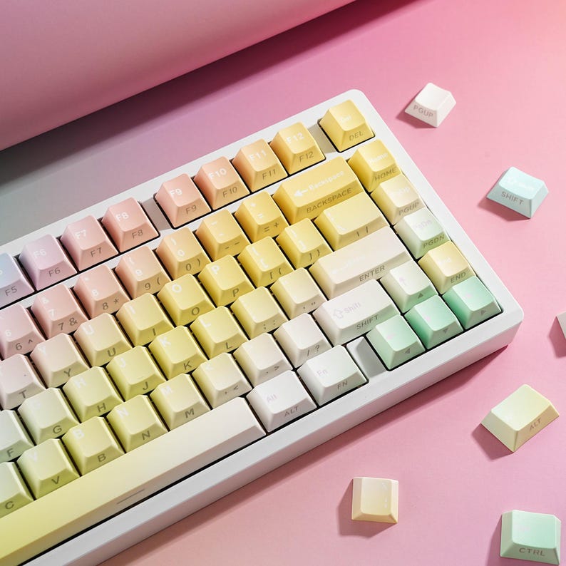 May include: A white mechanical keyboard with gradient-colored keycaps in pastel shades of pink, yellow, and green. Some keycaps are scattered around the keyboard on a pink surface. The keyboard has a compact layout.