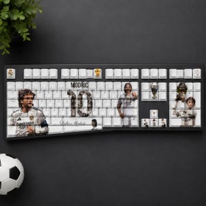 May include: A black computer keyboard with white keys featuring a custom graphic design of soccer player Luka Modric. The layout includes the text MODRIC, the number 10, and a signature, alongside images of the player in a white jersey and trophies.