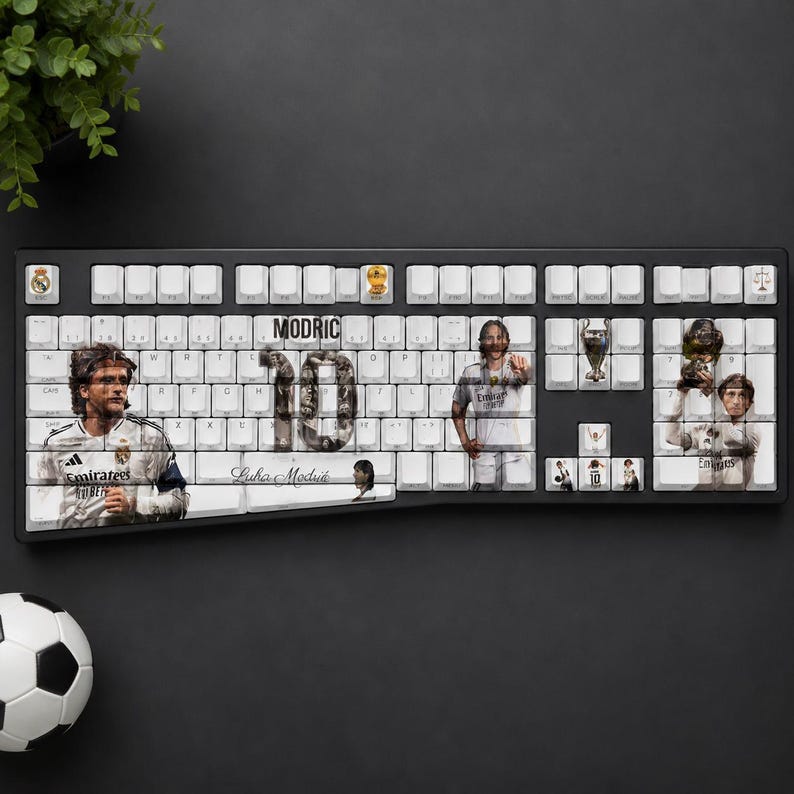 May include: A black computer keyboard with white keys featuring a custom graphic design of soccer player Luka Modric. The layout includes the text MODRIC, the number 10, and a signature, alongside images of the player in a white jersey and trophies.