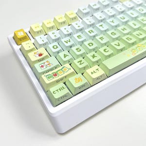 May include: A white mechanical keyboard with gradient keycaps transitioning from yellow to green. The keycaps feature standard letters, numbers, and symbols, along with custom illustrated keys with Chinese characters and cat illustrations.