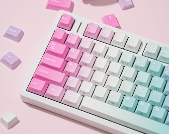 Puede incluir: Un teclado mecánico con un esquema de color degradado que transiciona de rosa a la izquierda, blanco en el centro y azul claro a la derecha. Las teclas tienen letras blancas y varias teclas sueltas en rosa y morado están esparcidas cerca.
