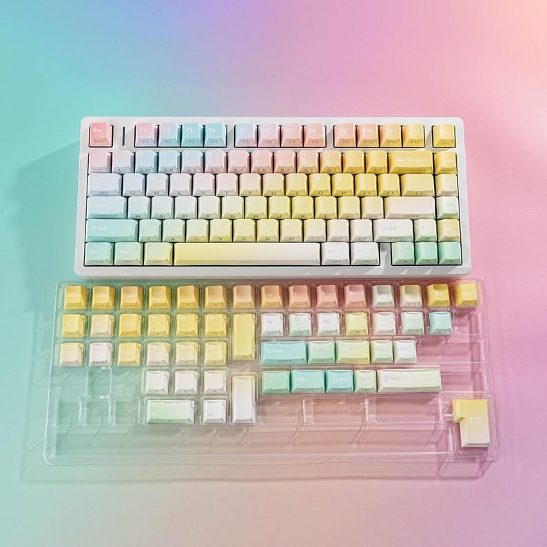 May include: A pastel-colored mechanical keyboard with a white frame and gradient keycaps in shades of pink, yellow, blue, and green. A clear plastic tray holds extra keycaps. The keyboard is designed for computer use.