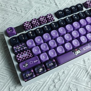 May include: A custom mechanical keyboard with purple and black keycaps. The keyboard features a mix of solid purple keys and patterned keys with a checkerboard design. Some keys have character illustrations.