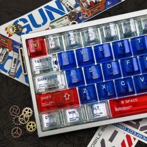 May include: A silver mechanical keyboard with red, clear, and blue keycaps. The keycaps are arranged in a standard QWERTY layout. The keyboard has a metal frame and includes decorative gears and text that reads "GUN".